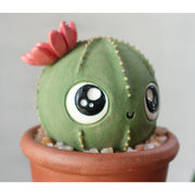 Ceramic sculpture of a round cactus with a red flower and very large cute kawaii eyes and a small smile. It is in a pot with rocks around it.