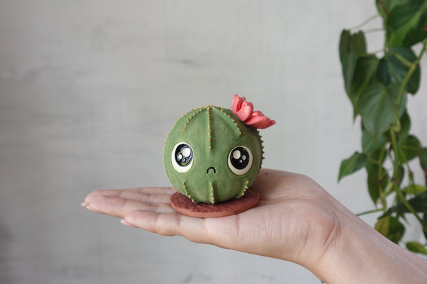 Ceramic sculpture of a round cactus with a red flower and very large cute kawaii eyes and a small frown. It is held in someone's hand.