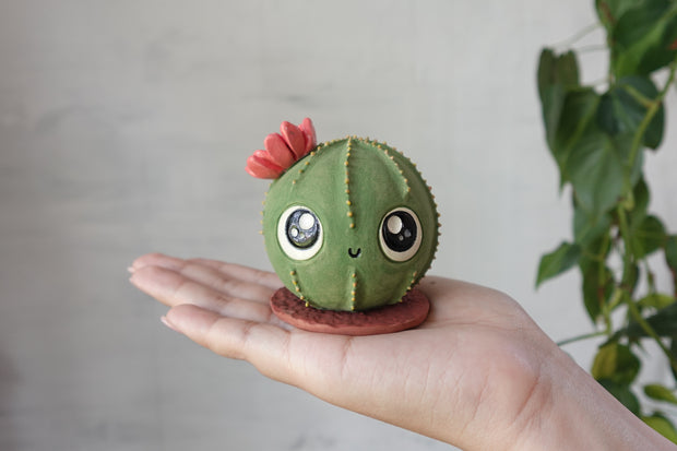 Ceramic sculpture of a round cactus with a red flower and very large cute kawaii eyes and a small smile. It is held in someone's hand.