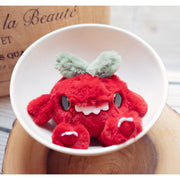 Small fluffy plush doll of a red tomato with large button eyes and a cute overbite smile. It has short arms and legs and a sage green leafy top.