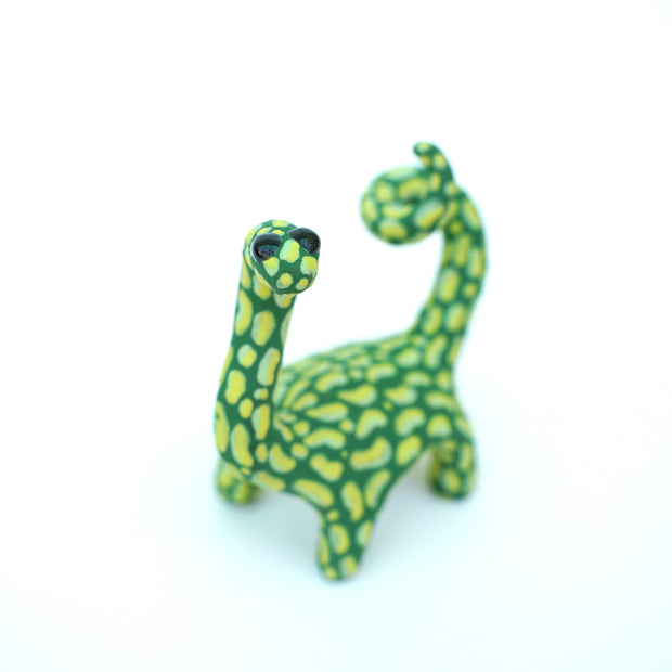 Ceramic sculpture of a green long neck dinosaur with large light green splotches as a pattern. It has a spiraled tail and black shiny eyes.