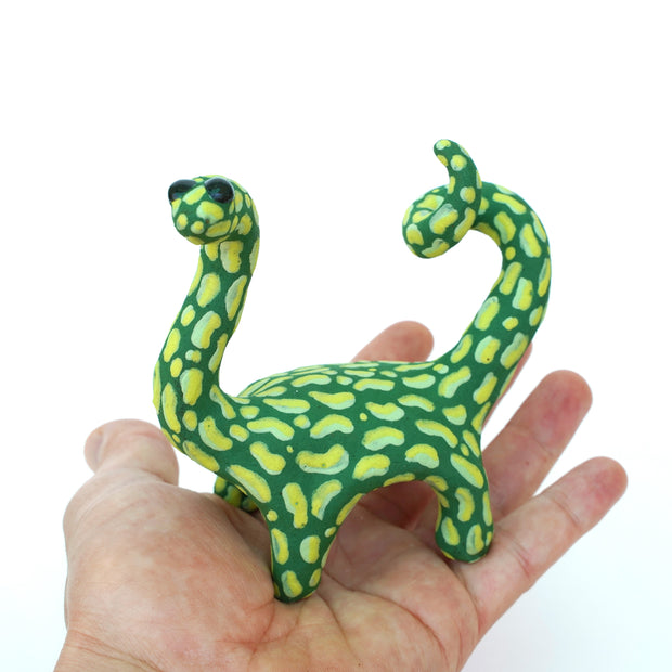 Ceramic sculpture of a green long neck dinosaur with large light green splotches as a pattern. It has a spiraled tail and black shiny eyes.