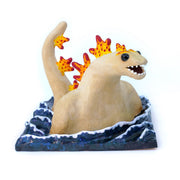 Ceramic sculpture of a tan Godzilla character, mostly submerged in waves of water which are depicted as a mostly flat square shaped base. Godzilla has flame colored spikes on its back and big black beads for eyes.