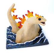 Ceramic sculpture of a tan Godzilla character, mostly submerged in waves of water which are depicted as a mostly flat square shaped base. Godzilla has flame colored spikes on its back and big black beads for eyes.