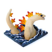 Ceramic sculpture of a tan Godzilla character, mostly submerged in waves of water which are depicted as a mostly flat square shaped base. Godzilla has flame colored spikes on its back and big black beads for eyes.