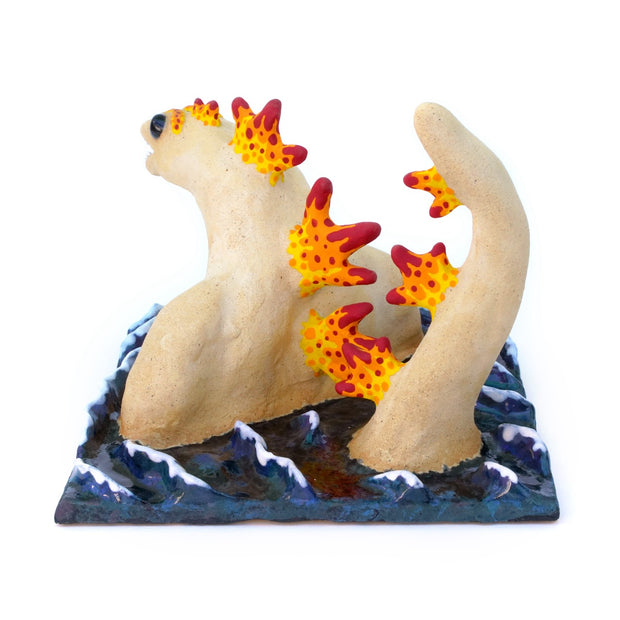 Ceramic sculpture of a tan Godzilla character, mostly submerged in waves of water which are depicted as a mostly flat square shaped base. Godzilla has flame colored spikes on its back and big black beads for eyes.