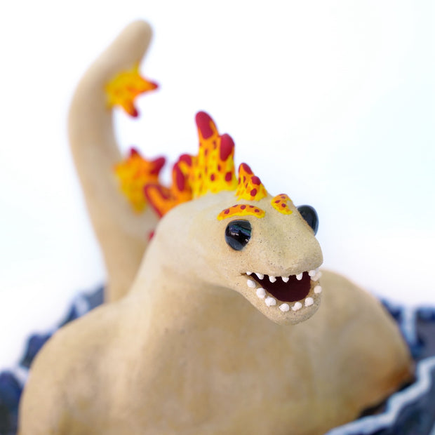 Ceramic sculpture of a tan Godzilla character, mostly submerged in waves of water which are depicted as a mostly flat square shaped base. Godzilla has flame colored spikes on its back and big black beads for eyes.