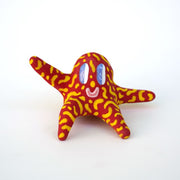 Small sculpture of a creature with large purple eyes and a smile, it is entirely red with a repeating yellow squiggle pattern. Its face takes up most of its body and it sits with long arms and legs extending out.