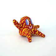 Small sculpture of a creature with large purple eyes and a smile, it is entirely red with a repeating yellow squiggle pattern. Its face takes up most of its body and it sits with long arms and legs extending out.