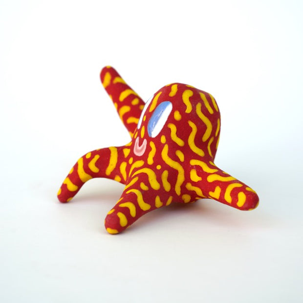 Small sculpture of a creature with large purple eyes and a smile, it is entirely red with a repeating yellow squiggle pattern. Its face takes up most of its body and it sits with long arms and legs extending out.