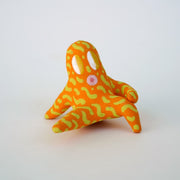 Small sculpture of a creature with large yellow eyes and an 'o' shaped mouth, it is entirely orange with a repeating green squiggle pattern. Its face takes up most of its body and it sits with long arms and legs extending out.