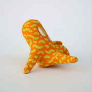 Small sculpture of a creature with large yellow eyes and an 'o' shaped mouth, it is entirely orange with a repeating green squiggle pattern. Its face takes up most of its body and it sits with long arms and legs extending out.