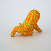 Small sculpture of a creature with large yellow eyes and an 'o' shaped mouth, it is entirely orange with a repeating green squiggle pattern. Its face takes up most of its body and it sits with long arms and legs extending out.