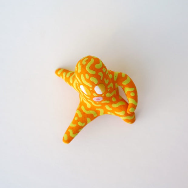 Small sculpture of a creature with large yellow eyes and an 'o' shaped mouth, it is entirely orange with a repeating green squiggle pattern. Its face takes up most of its body and it sits with long arms and legs extending out.