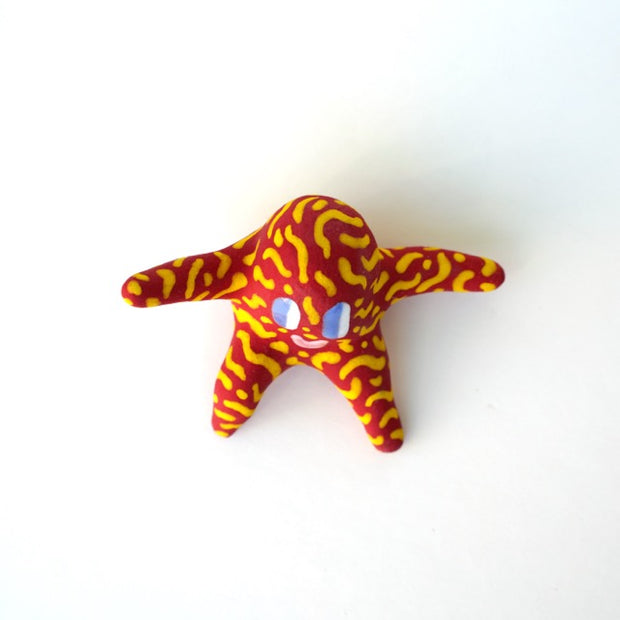 Small sculpture of a creature with large purple eyes and a smile, it is entirely red with a repeating yellow squiggle pattern. Its face takes up most of its body and it sits with long arms and legs extending out.