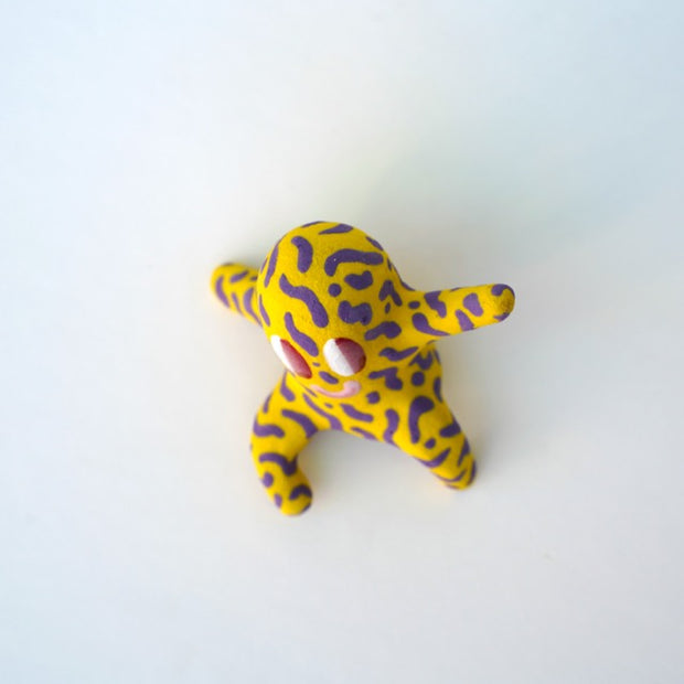 Small sculpture of a creature with large red eyes and a smile, it is entirely yellow with a repeating purple squiggle pattern. Its face takes up most of its body and it sits with long arms and legs extending out.