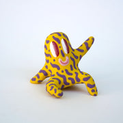 Small sculpture of a creature with large red eyes and a smile, it is entirely yellow with a repeating purple squiggle pattern. Its face takes up most of its body and it sits with long arms and legs extending out.