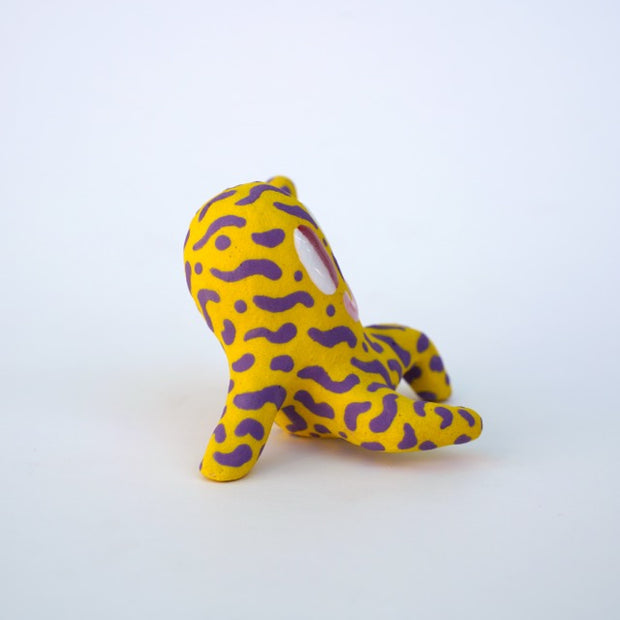 Small sculpture of a creature with large red eyes and a smile, it is entirely yellow with a repeating purple squiggle pattern. Its face takes up most of its body and it sits with long arms and legs extending out.