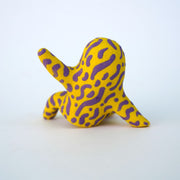 Small sculpture of a creature with large red eyes and a smile, it is entirely yellow with a repeating purple squiggle pattern. Its face takes up most of its body and it sits with long arms and legs extending out.