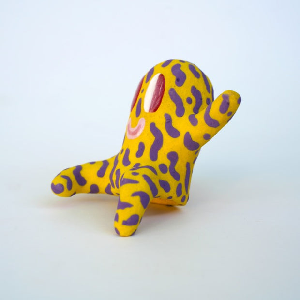 Small sculpture of a creature with large red eyes and a smile, it is entirely yellow with a repeating purple squiggle pattern. Its face takes up most of its body and it sits with long arms and legs extending out.