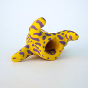 Small sculpture of a creature with large red eyes and a smile, it is entirely yellow with a repeating purple squiggle pattern. Its face takes up most of its body and it sits with long arms and legs extending out.