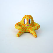 Small sculpture of a creature with large purple eyes and a surprised open mouth, it is entirely light green with a repeating orange squiggle pattern. Its face takes up most of its body and it sits with long arms and legs extending out.