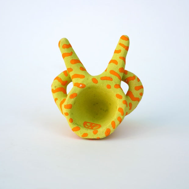 Small sculpture of a creature with large purple eyes and a surprised open mouth, it is entirely light green with a repeating orange squiggle pattern. Its face takes up most of its body and it sits with long arms and legs extending out.