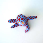 Small sculpture of a creature with large orange eyes and a smile, it is entirely blue with a repeating red squiggle pattern. Its face takes up most of its body and it sits with long arms and legs extending out.