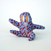 Small sculpture of a creature with large orange eyes and a smile, it is entirely blue with a repeating red squiggle pattern. Its face takes up most of its body and it sits with long arms and legs extending out.