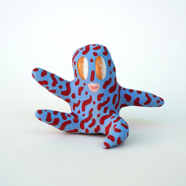 Small sculpture of a creature with large orange eyes and a smile, it is entirely blue with a repeating red squiggle pattern. Its face takes up most of its body and it sits with long arms and legs extending out.