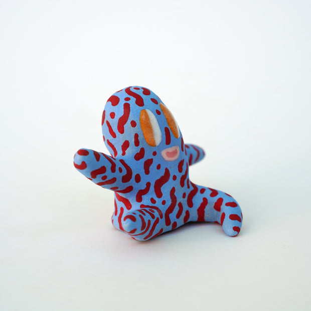 Small sculpture of a creature with large orange eyes and a smile, it is entirely blue with a repeating red squiggle pattern. Its face takes up most of its body and it sits with long arms and legs extending out.