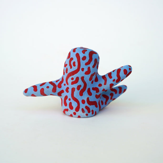 Small sculpture of a creature with large orange eyes and a smile, it is entirely blue with a repeating red squiggle pattern. Its face takes up most of its body and it sits with long arms and legs extending out.