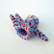 Small sculpture of a creature with large orange eyes and a smile, it is entirely blue with a repeating red squiggle pattern. Its face takes up most of its body and it sits with long arms and legs extending out.