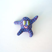 Small sculpture of a creature with large green eyes and a smile, it is entirely purple with a repeating blue squiggle pattern. Its face takes up most of its body and it leans back, balancing its weight with one arm down and its 2 legs. The other arm is extended up and out.