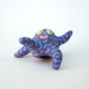 Small sculpture of a creature with large green eyes and a smile, it is entirely purple with a repeating blue squiggle pattern. Its face takes up most of its body and it leans back, balancing its weight with one arm down and its 2 legs. The other arm is extended up and out.