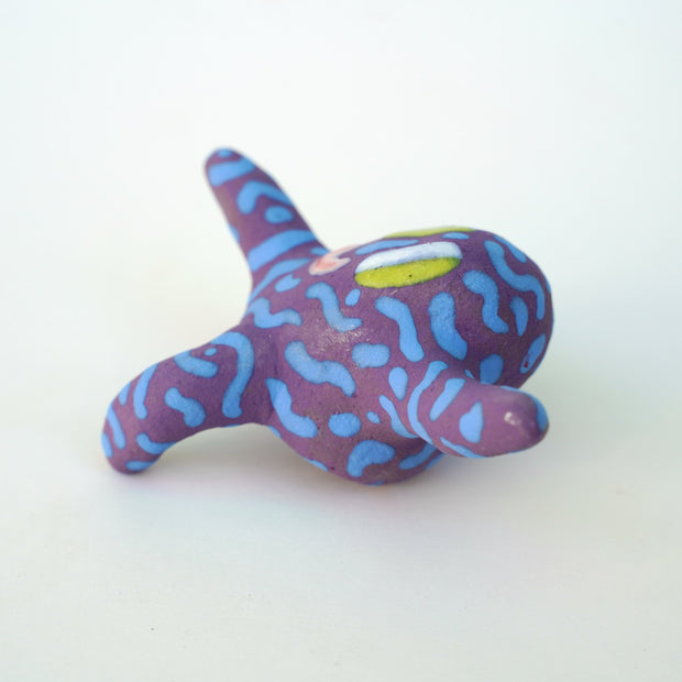 Small sculpture of a creature with large green eyes and a smile, it is entirely purple with a repeating blue squiggle pattern. Its face takes up most of its body and it leans back, balancing its weight with one arm down and its 2 legs. The other arm is extended up and out.