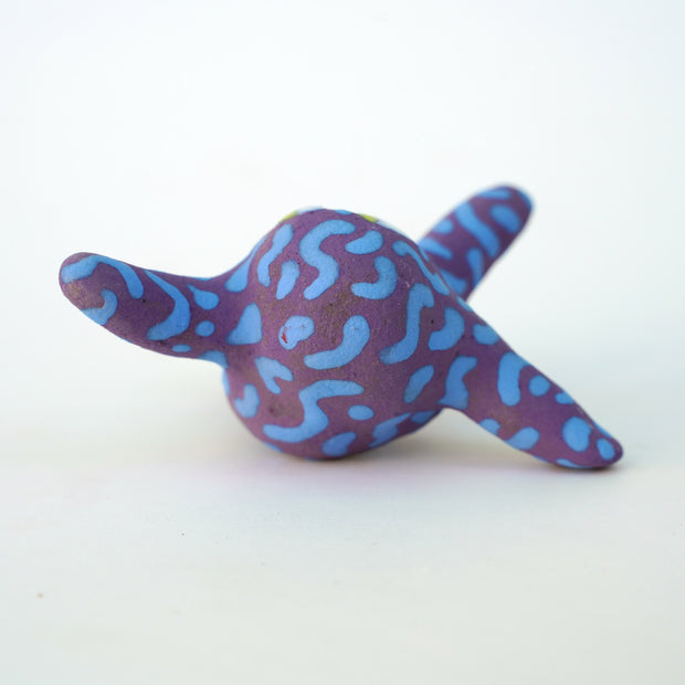 Small sculpture of a creature with large green eyes and a smile, it is entirely purple with a repeating blue squiggle pattern. Its face takes up most of its body and it leans back, balancing its weight with one arm down and its 2 legs. The other arm is extended up and out.