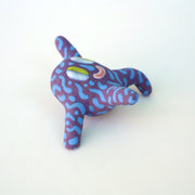 Small sculpture of a creature with large green eyes and a smile, it is entirely purple with a repeating blue squiggle pattern. Its face takes up most of its body and it leans back, balancing its weight with one arm down and its 2 legs. The other arm is extended up and out.