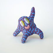 Small sculpture of a creature with large green eyes and a smile, it is entirely purple with a repeating blue squiggle pattern. Its face takes up most of its body and it leans back, balancing its weight with one arm down and its 2 legs. The other arm is extended up and out.