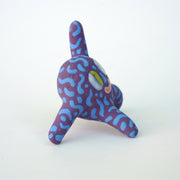 Small sculpture of a creature with large green eyes and a smile, it is entirely purple with a repeating blue squiggle pattern. Its face takes up most of its body and it leans back, balancing its weight with one arm down and its 2 legs. The other arm is extended up and out.