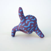 Small sculpture of a creature with large green eyes and a smile, it is entirely purple with a repeating blue squiggle pattern. Its face takes up most of its body and it leans back, balancing its weight with one arm down and its 2 legs. The other arm is extended up and out.