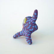 Small sculpture of a creature with large green eyes and a smile, it is entirely purple with a repeating blue squiggle pattern. Its face takes up most of its body and it leans back, balancing its weight with one arm down and its 2 legs. The other arm is extended up and out.