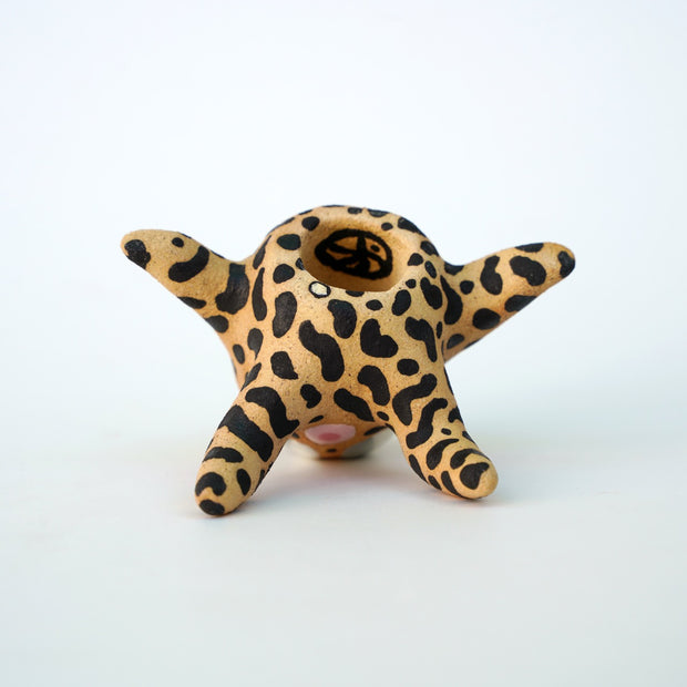 Small sculpture of a creature with large gray eyes and an 'o' shaped mouth, it is entirely brown with a repeating black squiggle pattern. Its face takes up most of its body and it leans back, on its 2 long arms and bends its long legs in front of it.