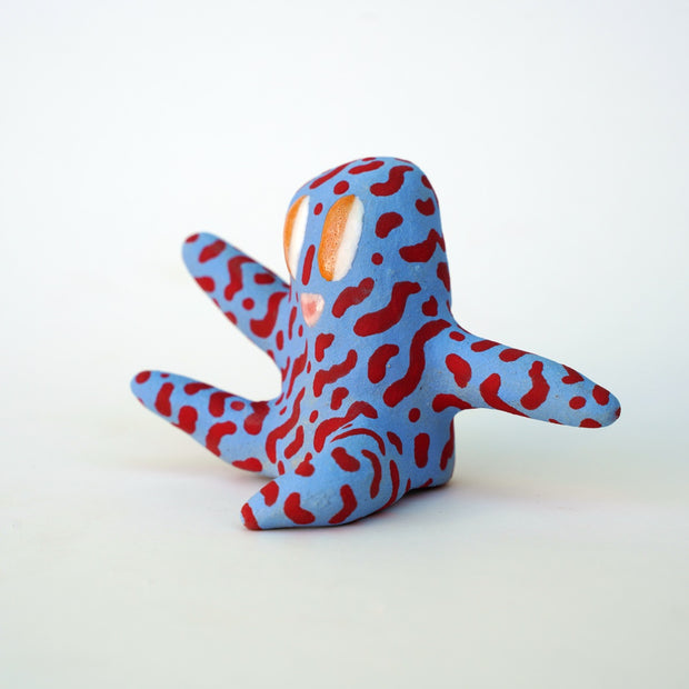 Small sculpture of a creature with large orange eyes and a smile, it is entirely blue with a repeating red squiggle pattern. Its face takes up most of its body and it sits with long arms and legs extending out.