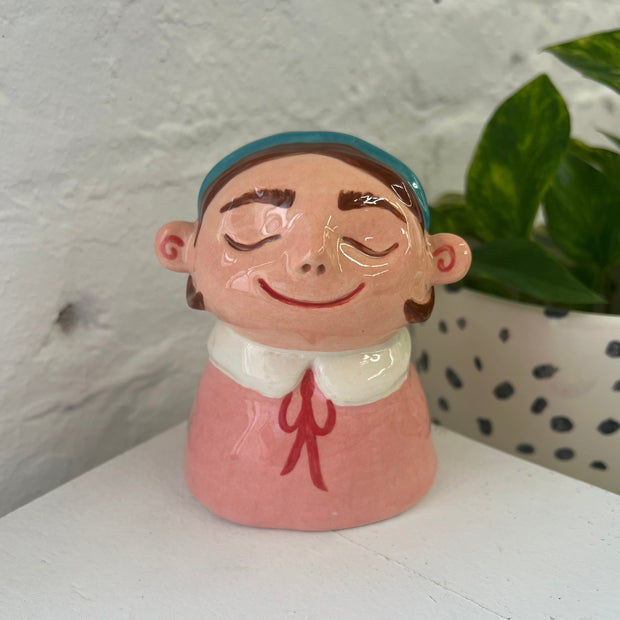 Ceramic bust of a cartoon style girl with brown hair and a teal headband. She has her eyes closed, smiling and wears a pink shirt with a white collar and red bow.