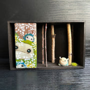 Horizontal open shadowbox art with a small sculpture of a blue horned monster, amongst tree branches and next to a painted panel with 3 similar monsters, stacked atop one another.