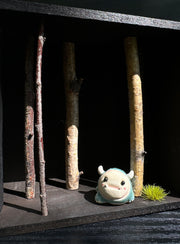 Horizontal open shadowbox art with a small sculpture of a blue horned monster, amongst tree branches and next to a painted panel with 3 similar monsters, stacked atop one another.