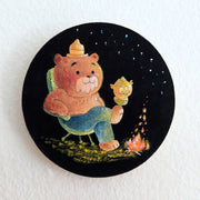 Illustration of a cute cartoon Smokey the Bear, sitting in front of a small campfire in a camping chair. He crosses his legs and Hooty the Owl rests on its foot. Stars light up the night sky.