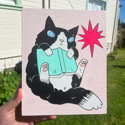 Painting on wood panel of a cartoon cat, fluffy with black and white fur. It holds a teal covered book against its chest and leans backwards, with its feet faced up. Background is pastel pink and a single bright pink starburst design element.