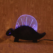 Wooden sculpture of a black dinosaur, flat mostly and emphasizing its silhouette.  Atop its back is a rounded plastic sheet with a line of lights, illuminating it and the interior of its mouth.